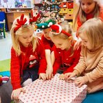 Dziewczynki w świątecznych opaskach mikołajkowych rozpakowują prezent przedszkolny od Świę.jpg