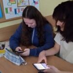 Zdjęcie 7-przedstawia dwie uczennice siedzące w ławce, trzymające telefony w dłoniach i uśmiechające się do siebie.