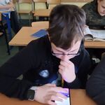 Zdjęcie 8- jeden chłopiec siedzi w ławce szkolnej, na ławce leży telefon. Chłopiec ma wyświetloną na telefonie klawiaturę. Z tyłu siedzi inny chłopiec w ławce szkolnej z telefonem w ręku i otwartą książką.