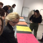 Nauczyciele w sali wykonują zadania warsztatowe pochylając się nad stołami na których leżą kolorowe kartki , w tle dwie tablice flipchart.