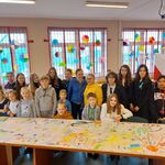 Na zdjęciu uczniowie SP12 w towarzystwie Pani Dyrektor Anny Satuła oraz nauczycieli ustawiają się do wspólnego zdjęcia grupowego. W tle okna ozdobione kolorowymi dekoracjami wykonanymi z kolorowej bibuły.