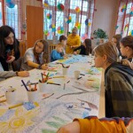 Uczniowie wykonują prace plastyczne wykorzystując technik 4K. Uczniowie siedzą przy stolikach malując na arkuszu papieru. Na stole kubeczki z kredkami.