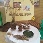 Na sztaludze jest gazetka na temat Tygodnia Zdrowego Chleba. Przed sztalugą stoi okrągły stół. Na stole znajdują się plecione  wazoniki (duży i mały) z kłosami zboża, koszyk z kromkami chleba, zielony talerz, wiązanka zbóż.