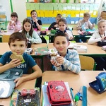 Dzieci z klasy drugiej siedzą w ławkach w klasie. Na stolikach leżą podręczniki i piórniki. Dzieci jedzą  kanapki z żytnim chlebem. 