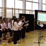 Dyrygentka szkolnego chóru wraz z uczniami ubranymi na galowo podczas śpiewania patriotycznych piosenek.