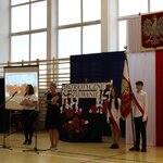 Poczet Sztandarowy na tle flagi Polski i godła podczas rozpoczęcia Patriotycznego Śpiewania. Po lewej stronie nauczycielka przy mikrofonie prowadząca uroczystość.