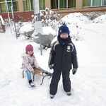 Chłopiec w czarnym kombinezonie ciągnie na sankach dziewczynkę z grupy Biedronki. W tle widać plac szkolny pokryty śniegiem..jpg