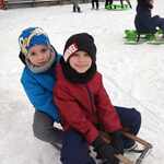 Dwóch chłopców ubranych w kombinezony zimowe siedzą na sankach. W tle widać inne dzieci bawiące się śniegiem..jpg